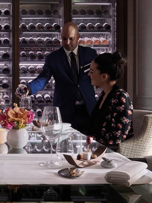 Man scans glass cellar as waitstaff pours wine in glass for woman at table