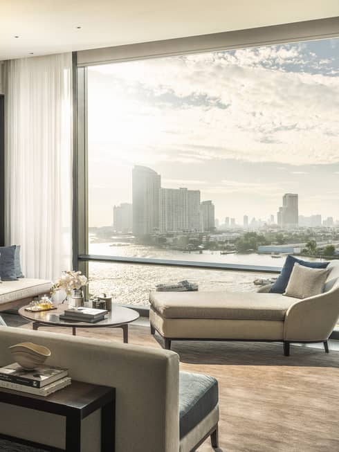 Hotel suite living area with neutral furniture, blue pillows, and large window overlooking a river and city skyline. An adjacent bedroom is visible with white bedding and flowers.