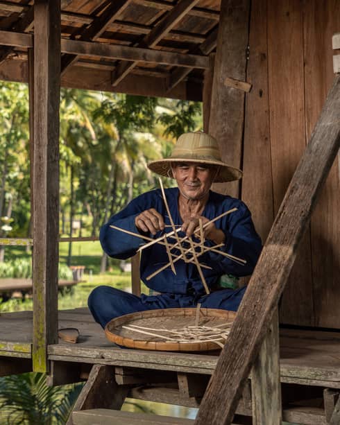 Man weaves a traditional Thai basket on the porch of a riverside pagoda