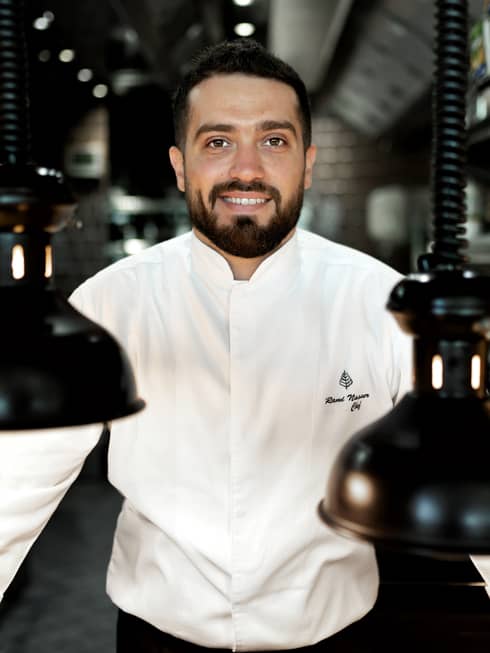 Chef Rami Nasser in his white chef's jacket smiles at the camera, flanked by black vintage pendant lamps hanging from coils.