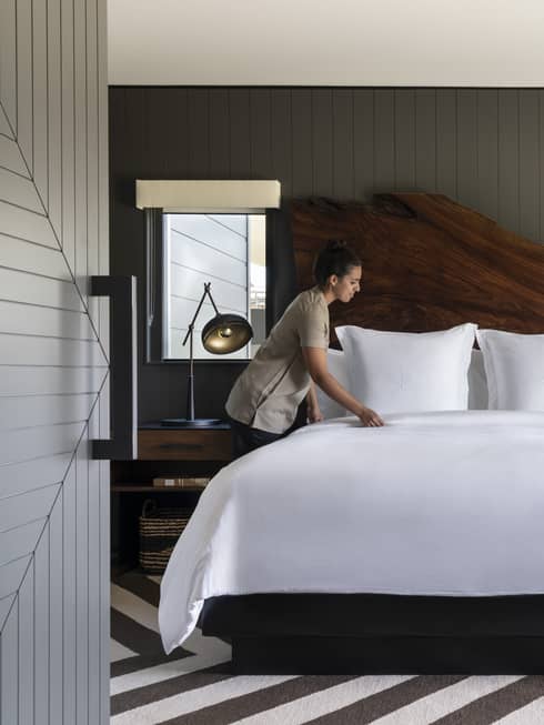 Four Seasons resort staff making bed with white linens in a luxury room featuring a wooden headboard, nightstands with lamps, striped rug and dark paneled walls
