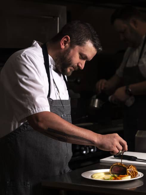A chef in a white shirt and a blue apron puts the finishing touches on several food dishes.