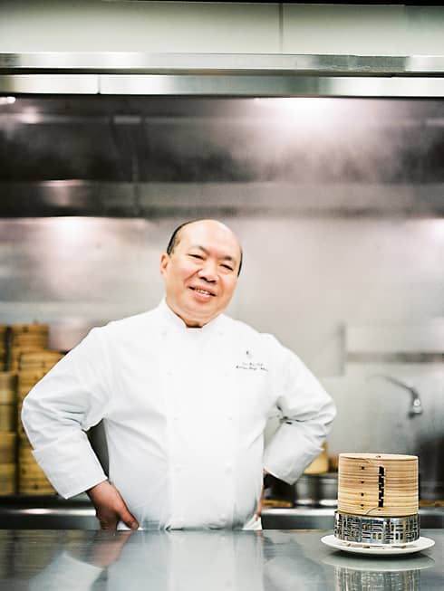 Chef Chan Yan Tak stands smiling in a professional kitchen with his hands on his hips, wearing a white uniform. Bamboo steamers are on the counter.