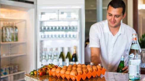 Smiling man holds green bottle in front of open fridge, fresh eggs on counter