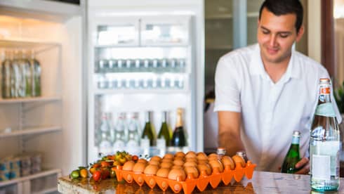 Smiling man holds green bottle in front of open fridge, fresh eggs on counter