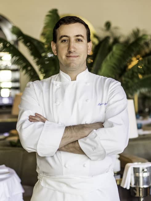 Chef Marco Calenzi stands in one of the Surfside restaurants at Four Seasons Surf Club. He is wearing a chef's coat and the restaurant has plants and white cloth tables.