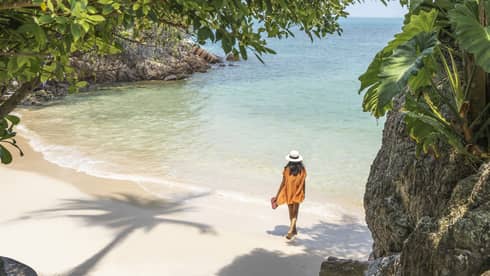 Woman in orange beach cover-up walks along the shoreline framed by a copse of native trees