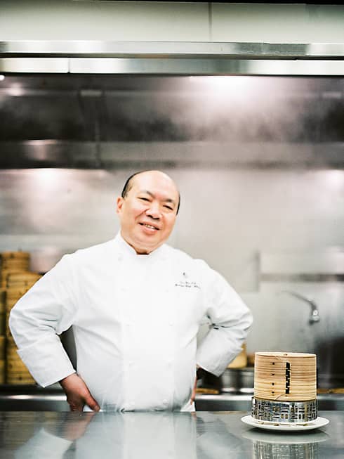 Chef Chan Yan Tak stands smiling in a professional kitchen with his hands on his hips, wearing a white uniform. Bamboo steamers are on the counter.