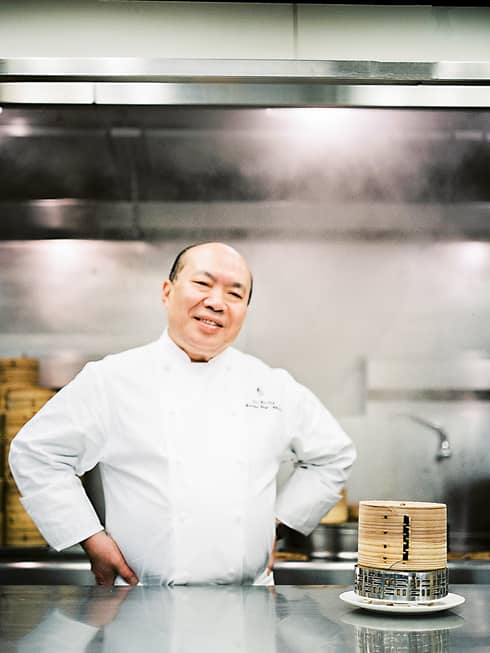 Chef Chan Yan Tak stands smiling in a professional kitchen with his hands on his hips, wearing a white uniform. Bamboo steamers are on the counter.