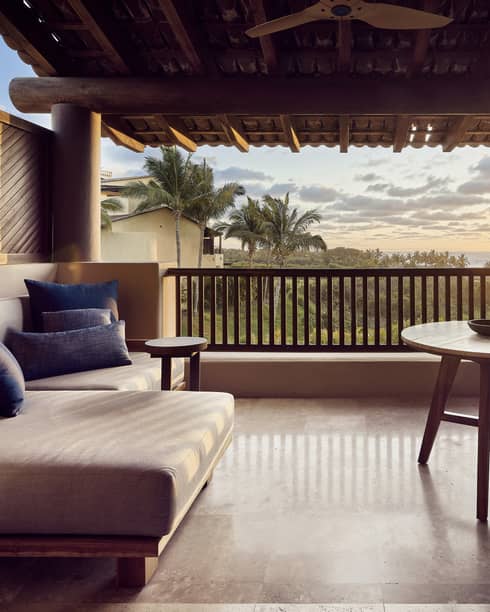 A sunlit hotel balcony with two chairs, a small table, and a sofa.
