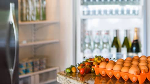 Smiling man holds green bottle in front of open fridge, fresh eggs on counter