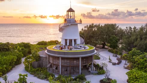 Lighthouse nestled among greenery with people on different levels, overlooking the ocean at sunset