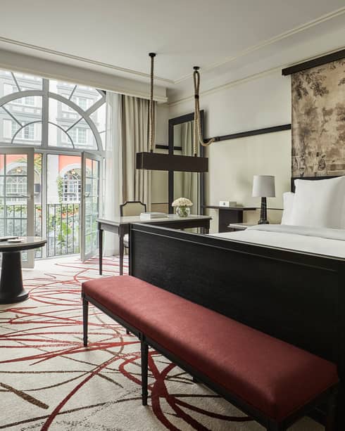 Premier Room bed under print headboard, red bench at foot, sunny arched balcony glass doors