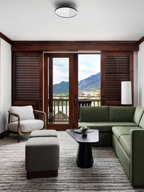 Bright sitting area with green sofa, armchair, two ottomans, coffee table, stone fireplace, TV and a balcony with mountain views through wooden shutters