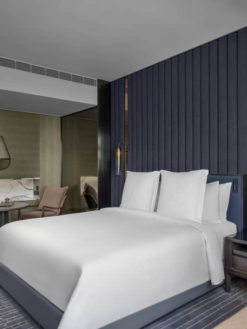 Renovated Deluxe City-View Room with bed and white linens, blue carpet and area rug, salmon chaise lounge, table for two with salmon armchairs