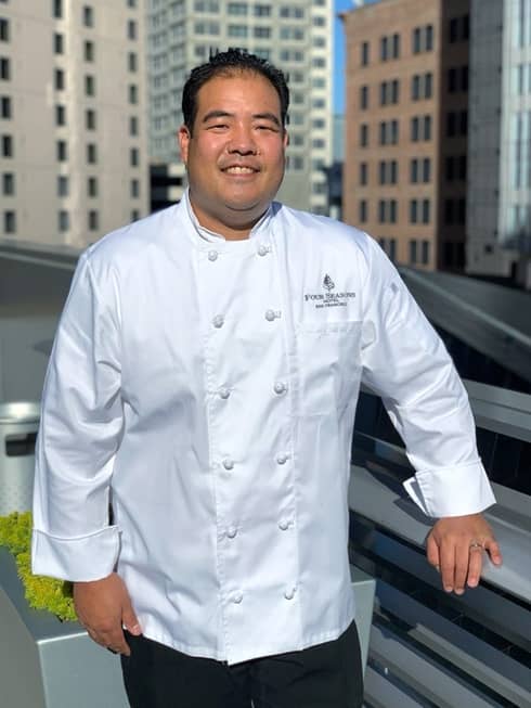 A male chef standing on a rooftop with a city behind him.