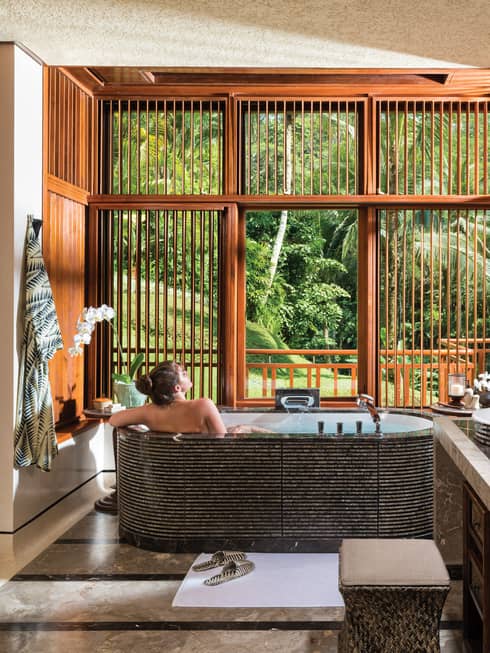 Woman with bare back relaxes in black freestanding spa tub near double sink, wood shutters open to lush garden view