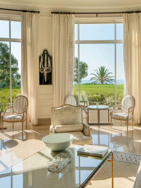 All-white salon with couches and dining chairs with tall rounded windows, lawn and sea view