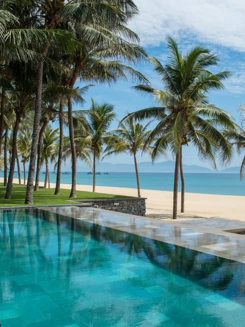 Tall palm trees between stone outdoor swimming pool and white sand beach