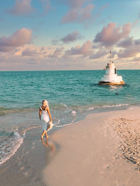 A woman walks along the shoreline towards a stone structure in the sea.