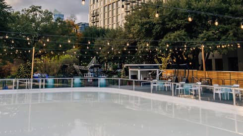 View of the Hotel overlooking an outdoor skating rink surrounded by trees and tables and lit by strings of Edison lights.