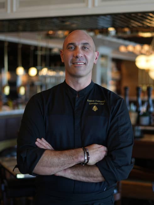 Person stands with arms crossed wearing a black chef's shirt with a restaurant dining room in the background