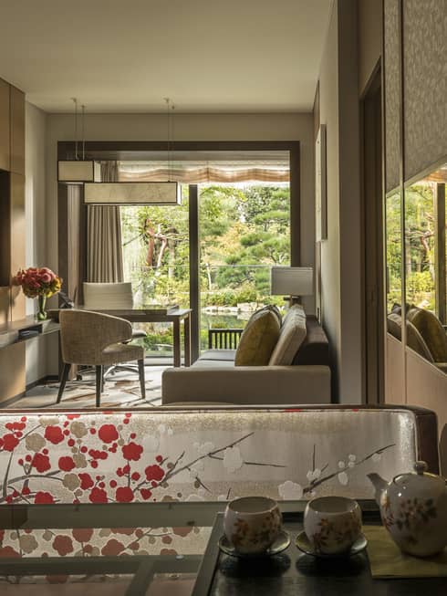 Connecting hotel room with sofa, desk and chairs, floral-patterned screen, tea set and large window overlooking a tranquil pond garden