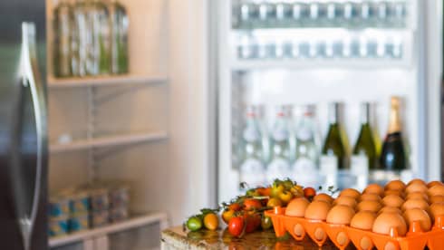Smiling man holds green bottle in front of open fridge, fresh eggs on counter