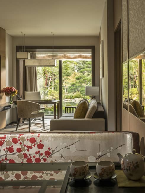 Connecting hotel room with sofa, desk and chairs, floral-patterned screen, tea set and large window overlooking a tranquil pond garden