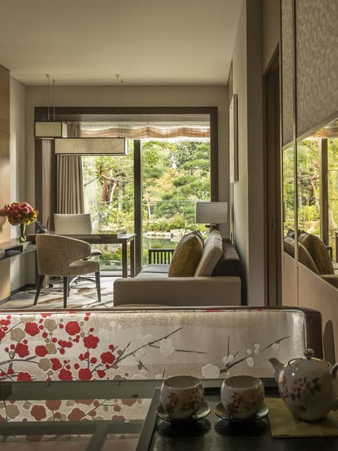 Connecting hotel room with sofa, desk and chairs, floral-patterned screen, tea set and large window overlooking a tranquil pond garden