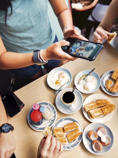 An aerial view of four friends eating breakfast while one of them captures an overhead image of the food on the table with her iphone