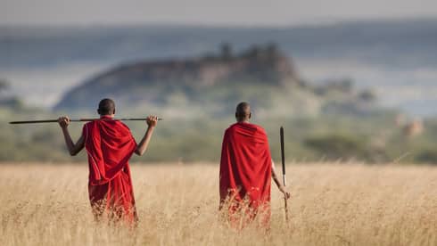 Two men in red indigenous clothing look out at a mountain on a savannah.