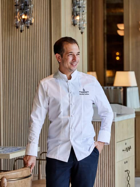 Chef with short dark hair wears a long-sleeve white chef's jacket and dark blue pants while standing in a dining room with light-wood furnishings