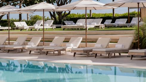 Luxury hotel outdoor pool area with rows of white lounge chairs, umbrellas and neatly landscaped greenery in the background