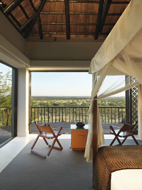 Poster bed, canopy by fold-up chairs, wood table by open wall with Serengeti views
