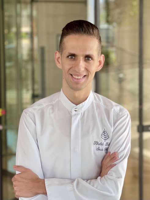 Man with short hair wearing white chef's jacket stands with arms crossed smiling at the camera