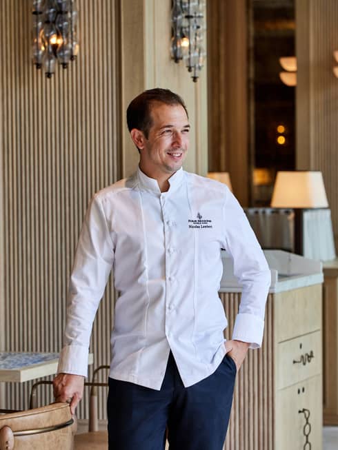 Chef with short dark hair wears a long-sleeve white chef's jacket and dark blue pants while standing in a dining room with light-wood furnishings