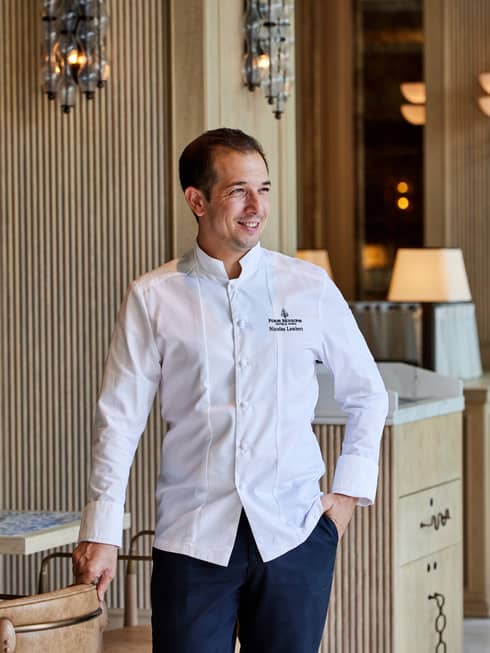 Chef with short dark hair wears a long-sleeve white chef's jacket and dark blue pants while standing in a dining room with light-wood furnishings