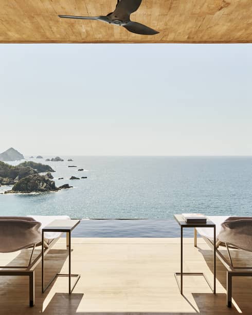 Open terrace with two lounge chairs and side tables, overlooking an infinity pool and a stunning ocean view with rocky islands. The space has a natural wood ceiling and serene ambience.