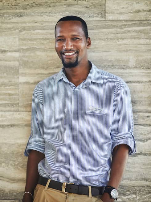 Smiling person in a checkered shirt standing against a stone wall indoors.