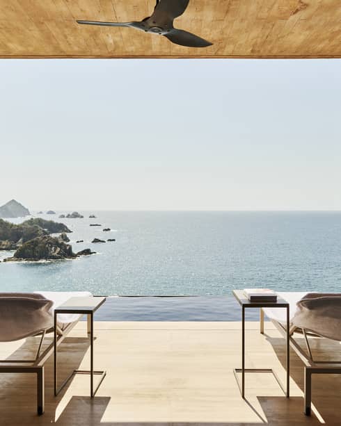 Open terrace with two lounge chairs and side tables, overlooking an infinity pool and a stunning ocean view with rocky islands. The space has a natural wood ceiling and serene ambience.