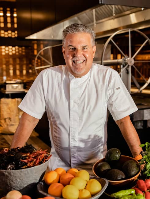 A smiling chef in white chefs clothes standing behind bowls of fruit and vegetables.