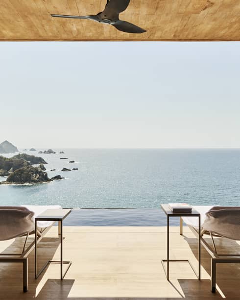 Open terrace with two lounge chairs and side tables, overlooking an infinity pool and a stunning ocean view with rocky islands. The space has a natural wood ceiling and serene ambience.