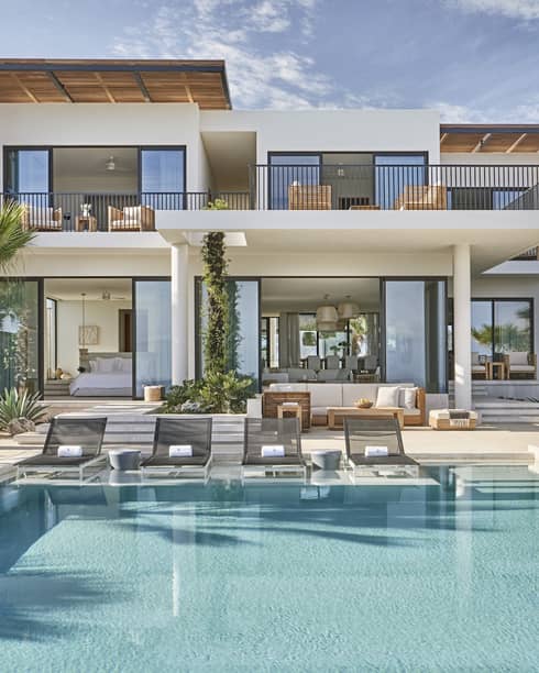 Modern luxury villa with a spacious pool area, surrounded by palm trees, featuring outdoor lounge chairs, a dining area, and floor-to-ceiling windows.