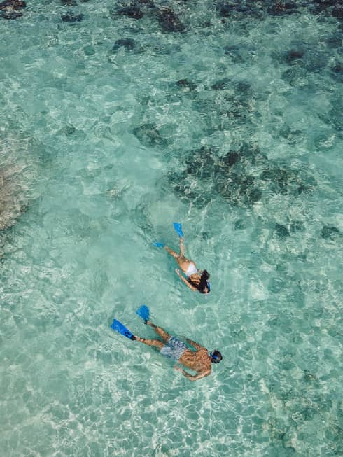 Aerial view of two snorkellers wearing blue flippers in an expanse of shallow, turquoise waters rippling over the reef.
