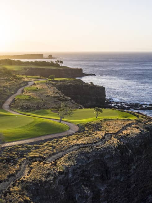 Soft sunlight filters over a pristine golf course with a winding road set on the edge of a cliff overlooking the calm ocean.