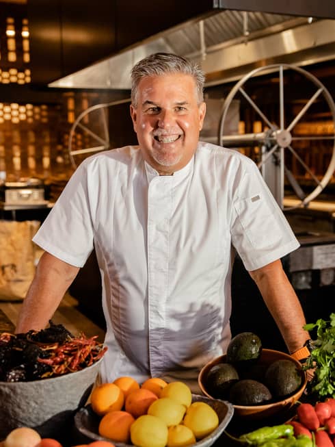 A smiling chef in white chefs clothes standing behind bowls of fruit and vegetables.