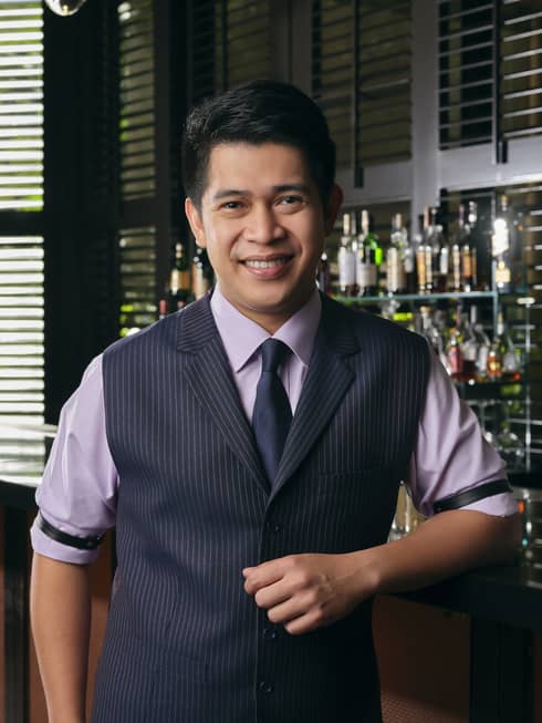 Smiling Bar Manager Gabriel Carlos poses for the camera, an artful display of liquor bottles on the shelf behind him