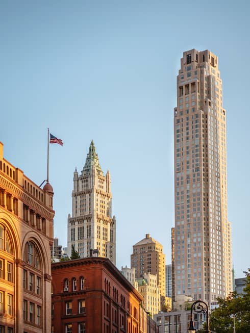 Downtown cityscape view featuring historic and modern skyscrapers, including the Woolworth Building, with an American flag against a clear blue sky
