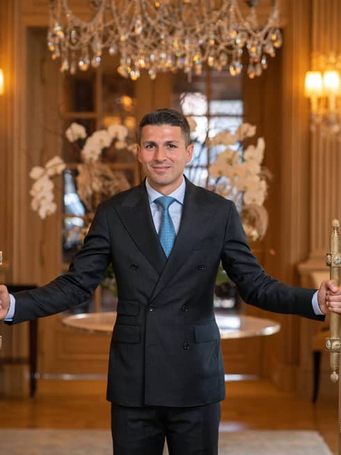 Salvatore Salerno in dark suit, standing and holding open French doors under chandelier